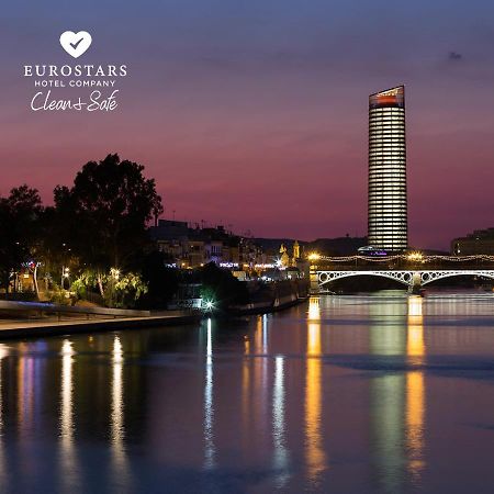Eurostars Torre Sevilla Exterior foto The photo depicts a serene riverside view at dusk. A tall, modern skyscraper with illuminated windows stands in the background against a twilight sky transitioning from orange to deep blue. On the left, there are trees and buildings that add to the u
