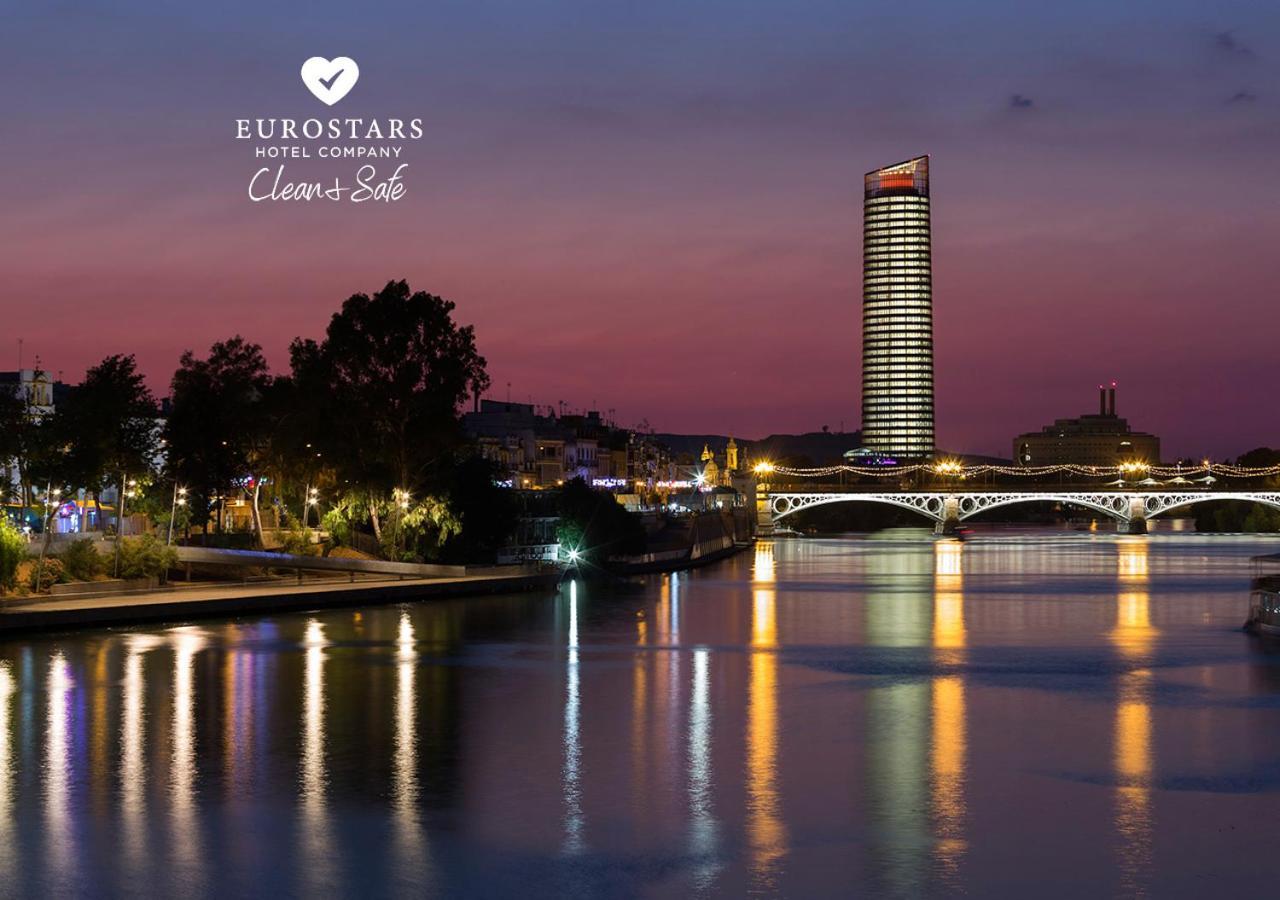 Eurostars Torre Sevilla Exterior foto The photo depicts a serene riverside view at dusk. A tall, modern skyscraper with illuminated windows stands in the background against a twilight sky transitioning from orange to deep blue. On the left, there are trees and buildings that add to the u