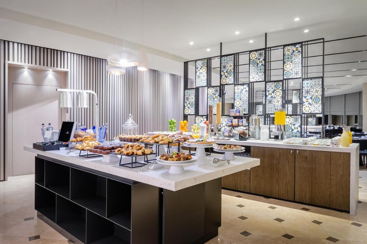 Eurostars Torre Sevilla Exterior foto The photo shows a modern buffet setup in a dining area. There is a stylish display featuring various food items, including pastries and desserts, arranged on a central island with multiple tiers. Behind the food spread, there is a decorative partitio