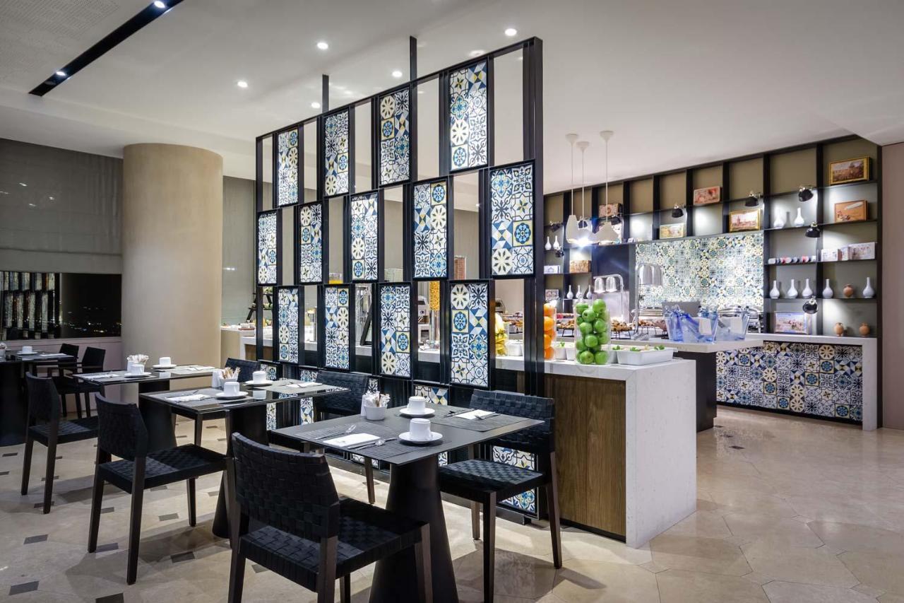 Eurostars Torre Sevilla Exterior foto The photo shows a restaurant interior featuring a modern design. There are several dining tables set for meal service, each with black chairs and neatly arranged tableware, including cups and plates. A decorative partition with colorful tile work sep