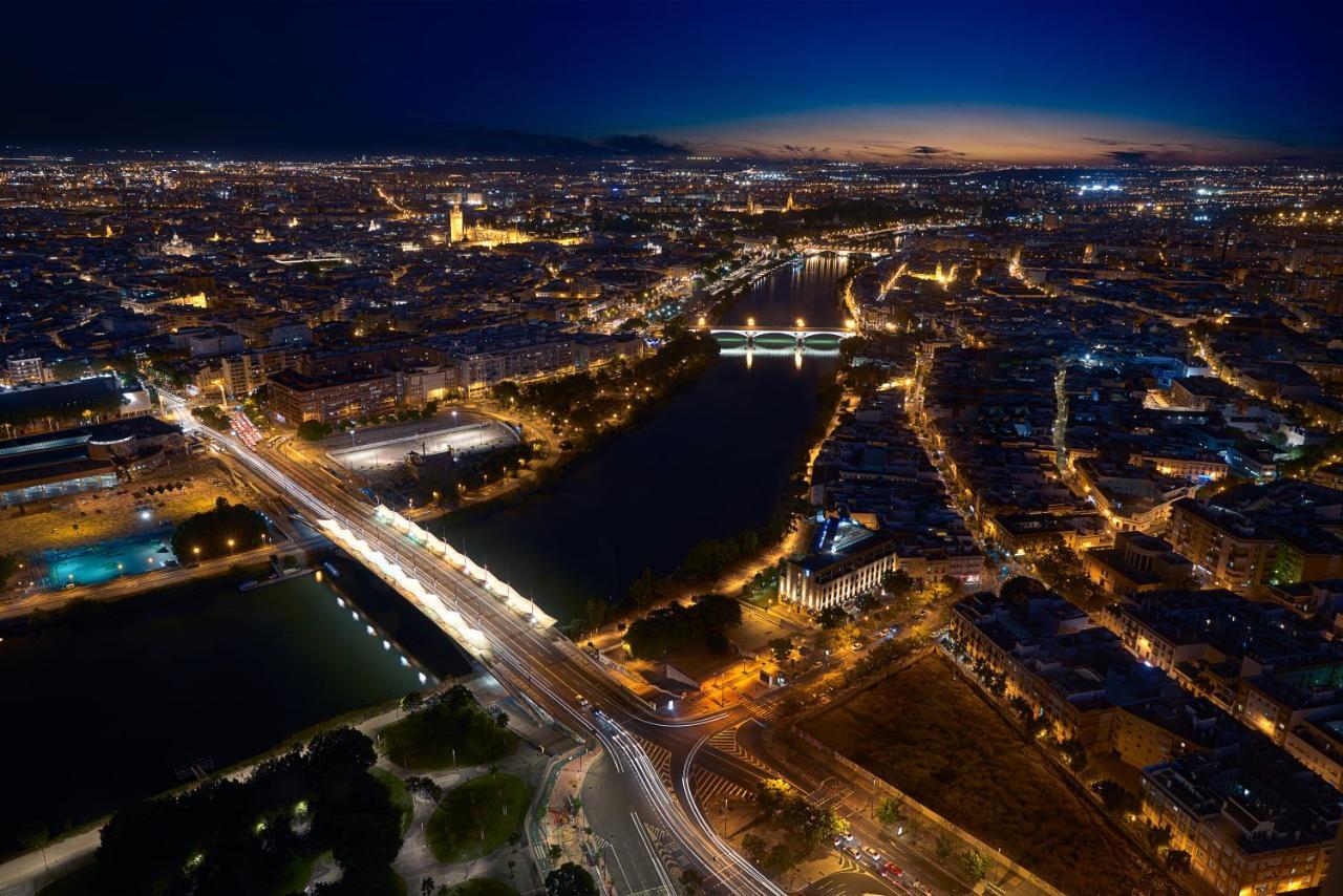 Eurostars Torre Sevilla Exterior foto The photo shows a panoramic view of a city at dusk, with a blend of natural and artificial light illuminating the landscape. A river runs through the scene, reflecting the city lights. There are several bridges crossing the river, and the buildings a