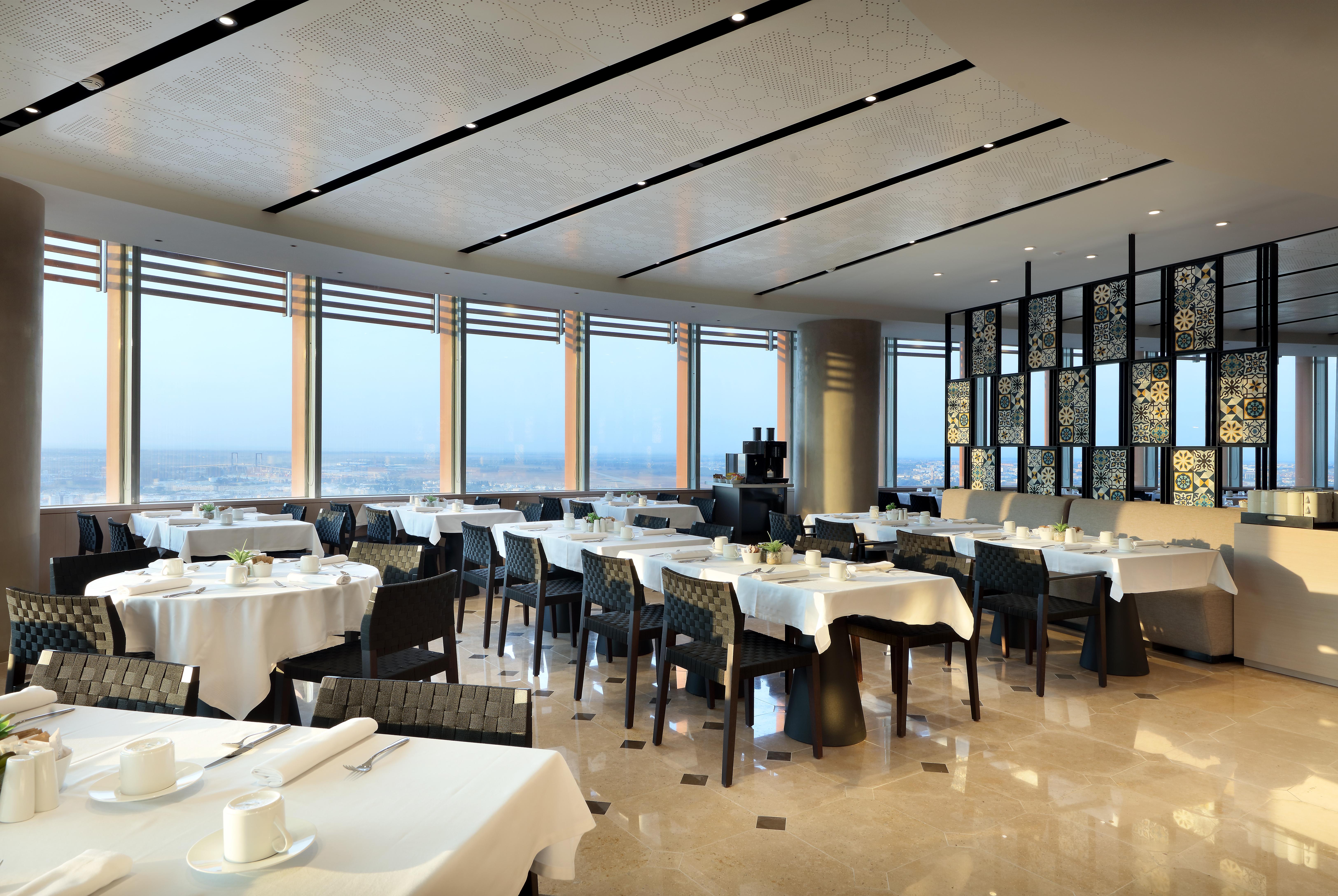 Eurostars Torre Sevilla Exterior foto The photo shows the interior of a modern restaurant. The dining area features several tables covered with white tablecloths, each set with utensils and cups. There are black chairs around the tables. The space is well-lit by large windows that likely