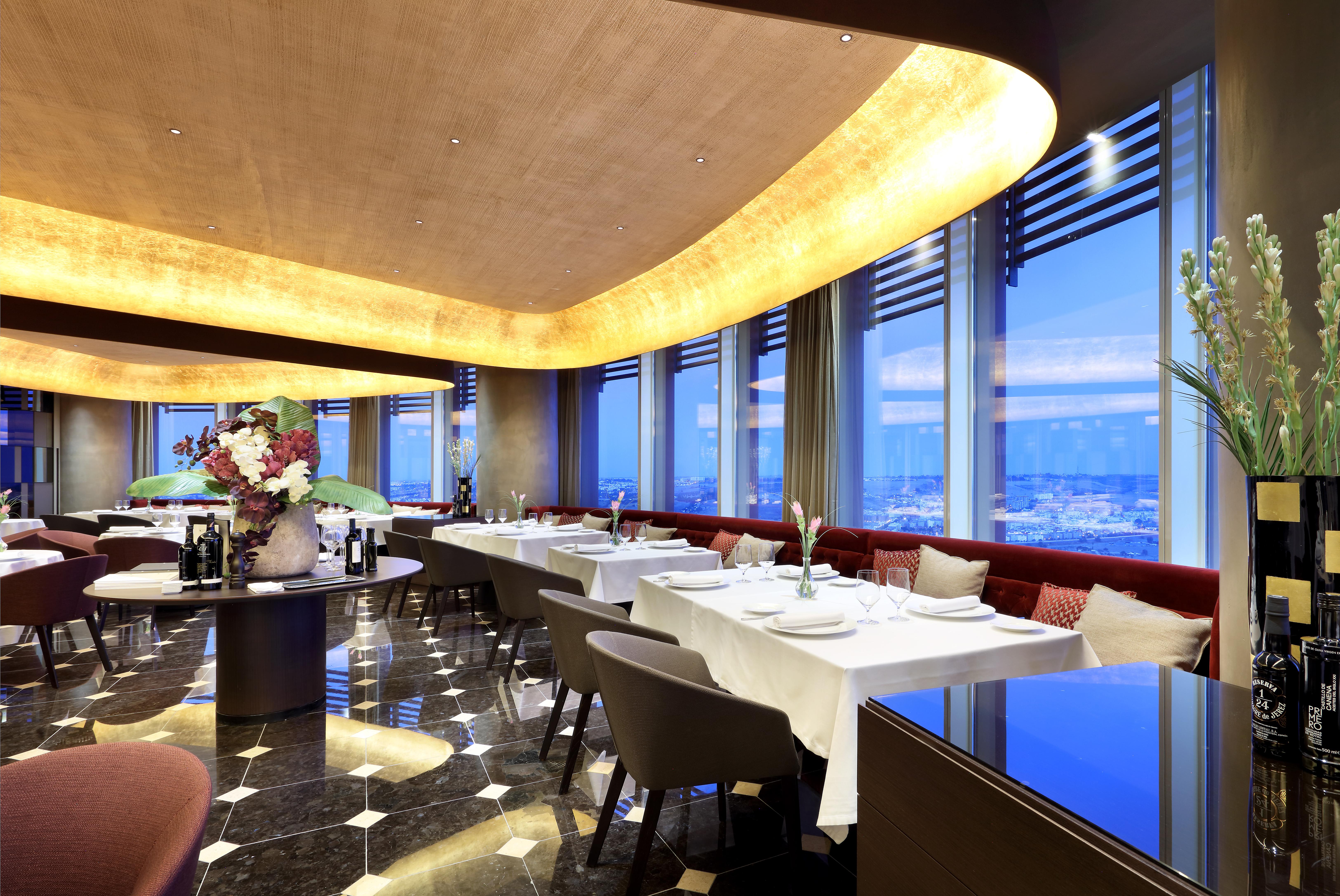 Eurostars Torre Sevilla Exterior foto The photo depicts an elegant restaurant interior with a modern design. The space features large windows that offer a panoramic view, likely showcasing a city skyline. The ceiling is curved and includes warm lighting that adds a cozy ambiance. There a