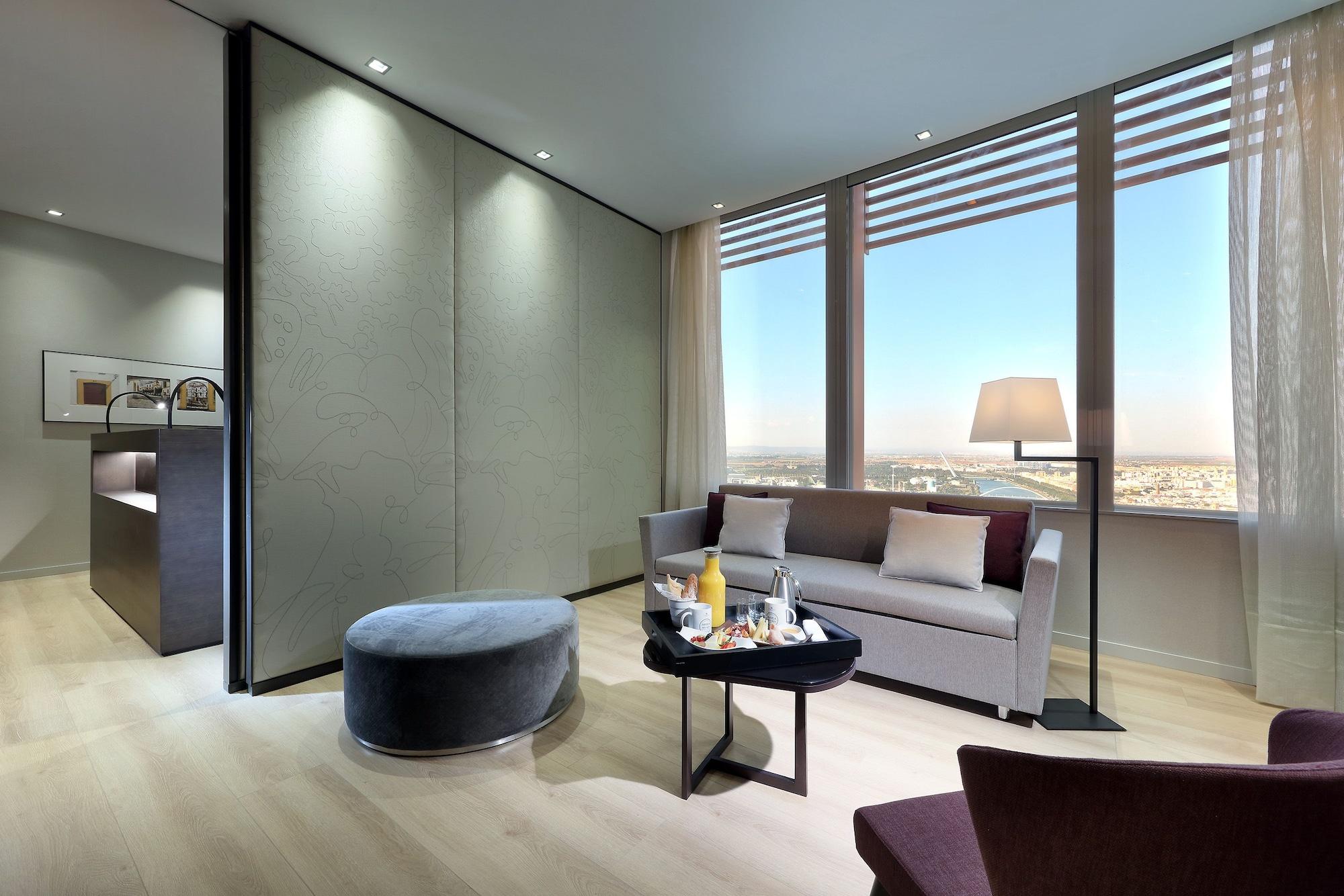 Eurostars Torre Sevilla Exterior foto The photo depicts a modern, stylish interior of a hotel room or suite. It features a comfortable seating area with a grey sofa, a round ottoman, and a small table that appears to hold refreshments. Large windows allow natural light to fill the space,