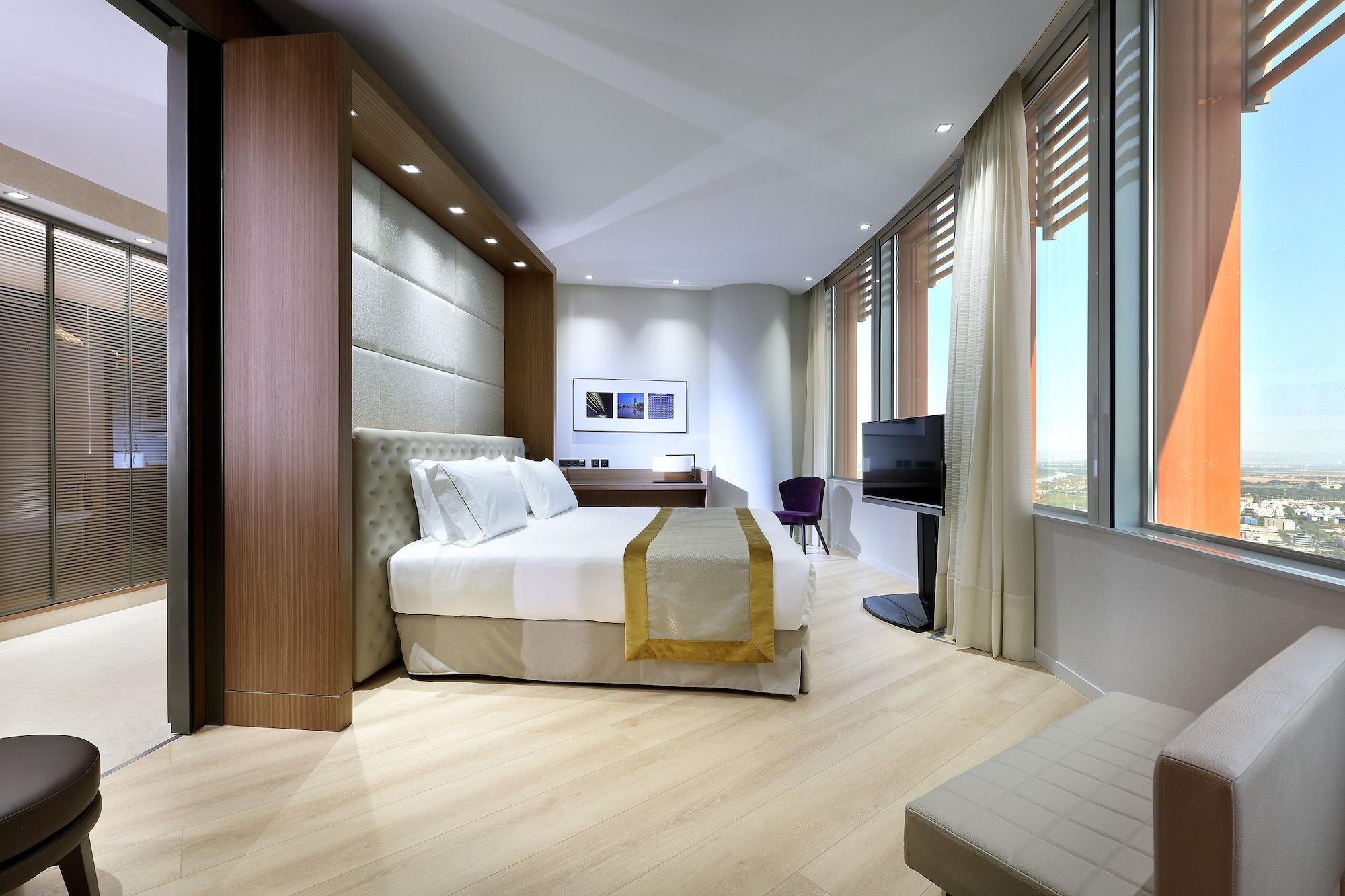 Eurostars Torre Sevilla Exterior foto The photo shows a modern hotel room featuring a large bed with a light-colored headboard. The decor is minimalist, with a neutral color palette and wooden accents. A small seating area is located next to the bed, and there is a desk with a chair. A t