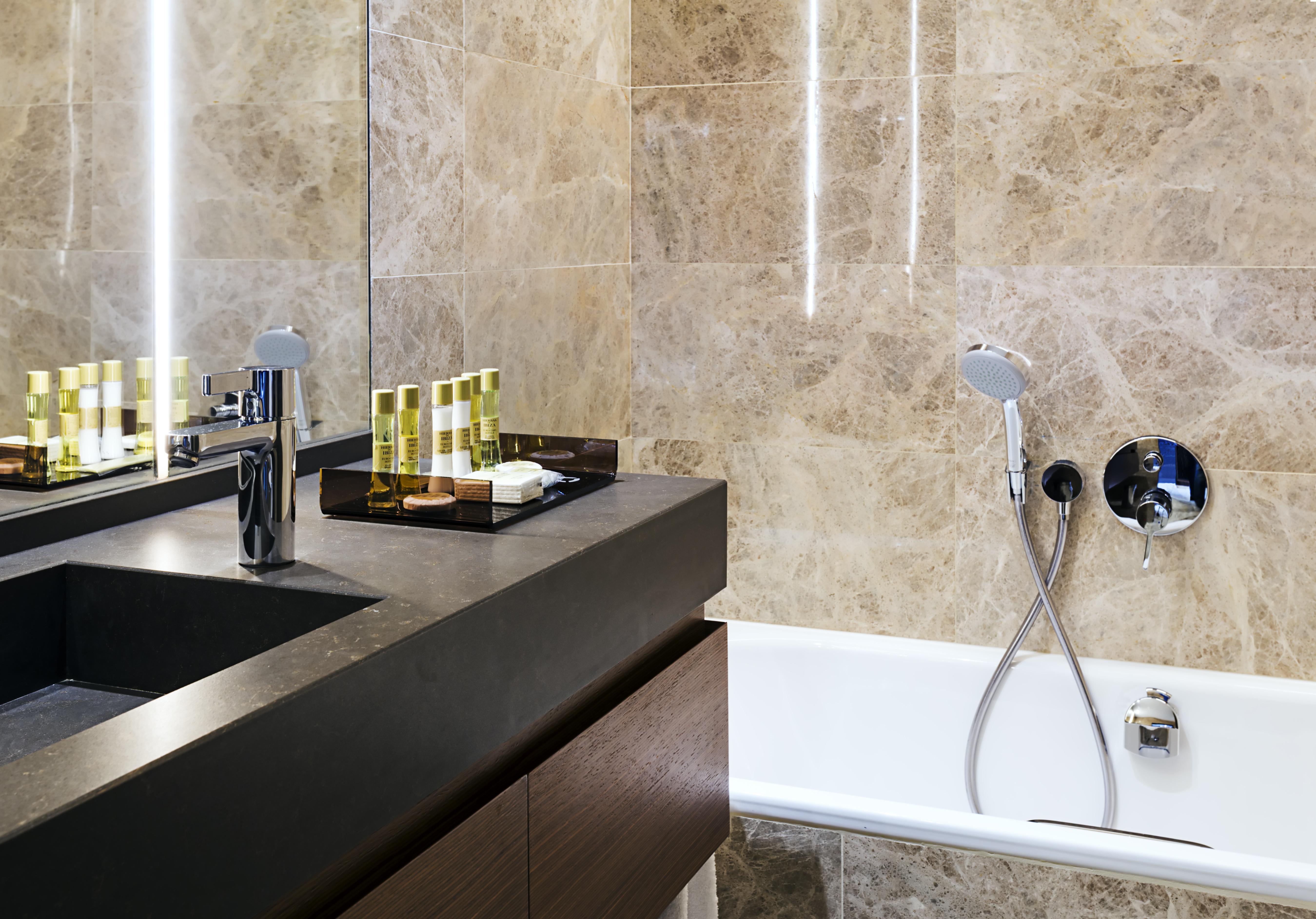 Eurostars Torre Sevilla Exterior foto The photo shows a modern bathroom featuring elegant design elements. There is a sleek, dark countertop with a rectangular sink and a modern faucet. On the countertop, there are several small bottles, likely containing toiletries, arranged neatly on a