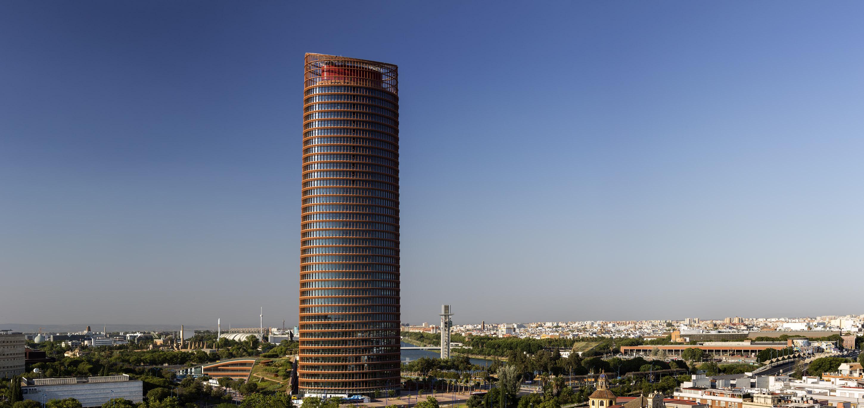 Eurostars Torre Sevilla Exterior foto The photo showcases a tall, modern skyscraper with a cylindrical shape and a distinctive facade featuring a combination of glass and metal elements. The building is surrounded by greenery and urban landscapes, with other structures visible in the bac