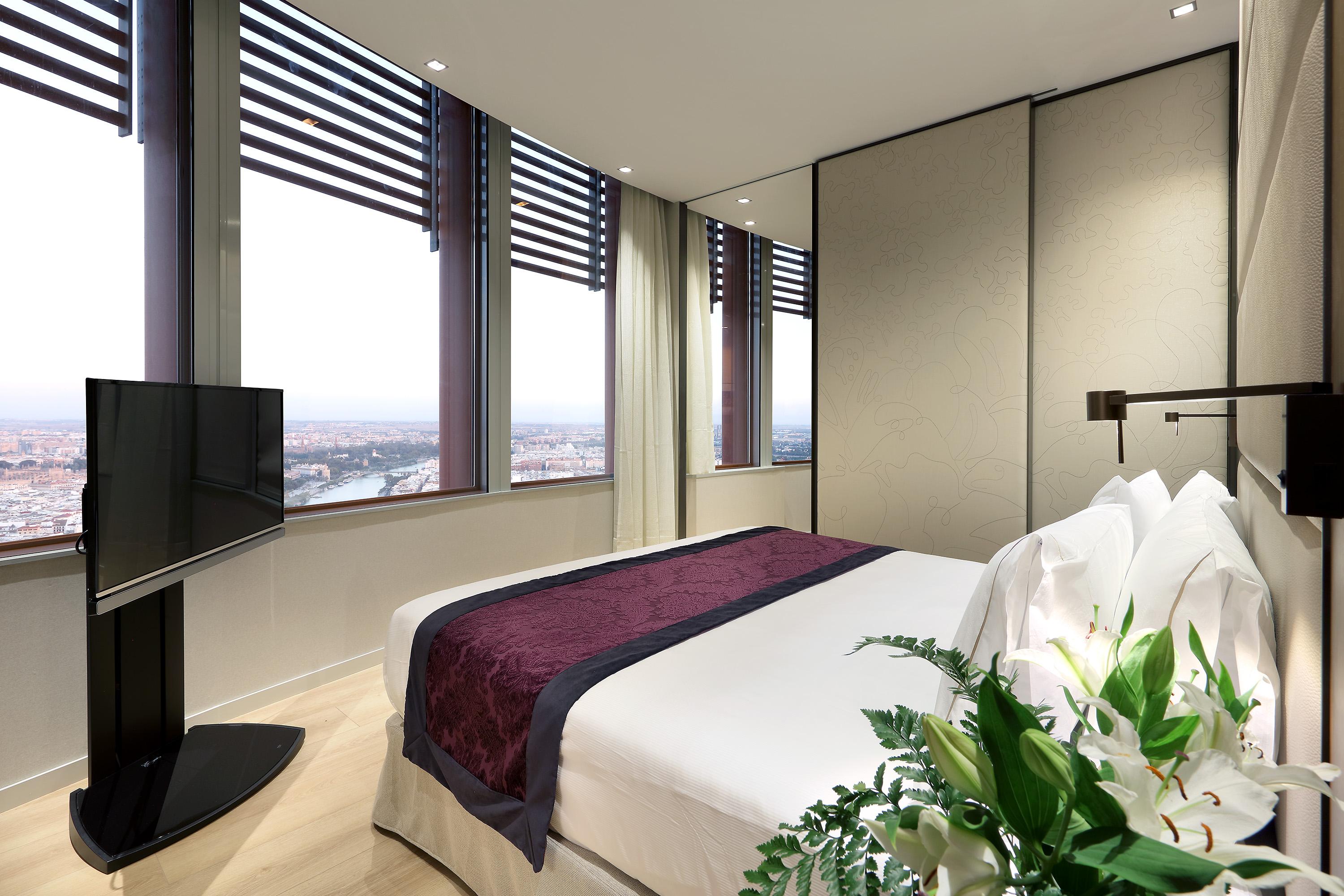 Eurostars Torre Sevilla Exterior foto The image shows a modern hotel room featuring a large bed with a dark purple bedspread and white pillows. The room has floor-to-ceiling windows with horizontal blinds, providing a panoramic view of the city outside. A flat-screen television is mounte