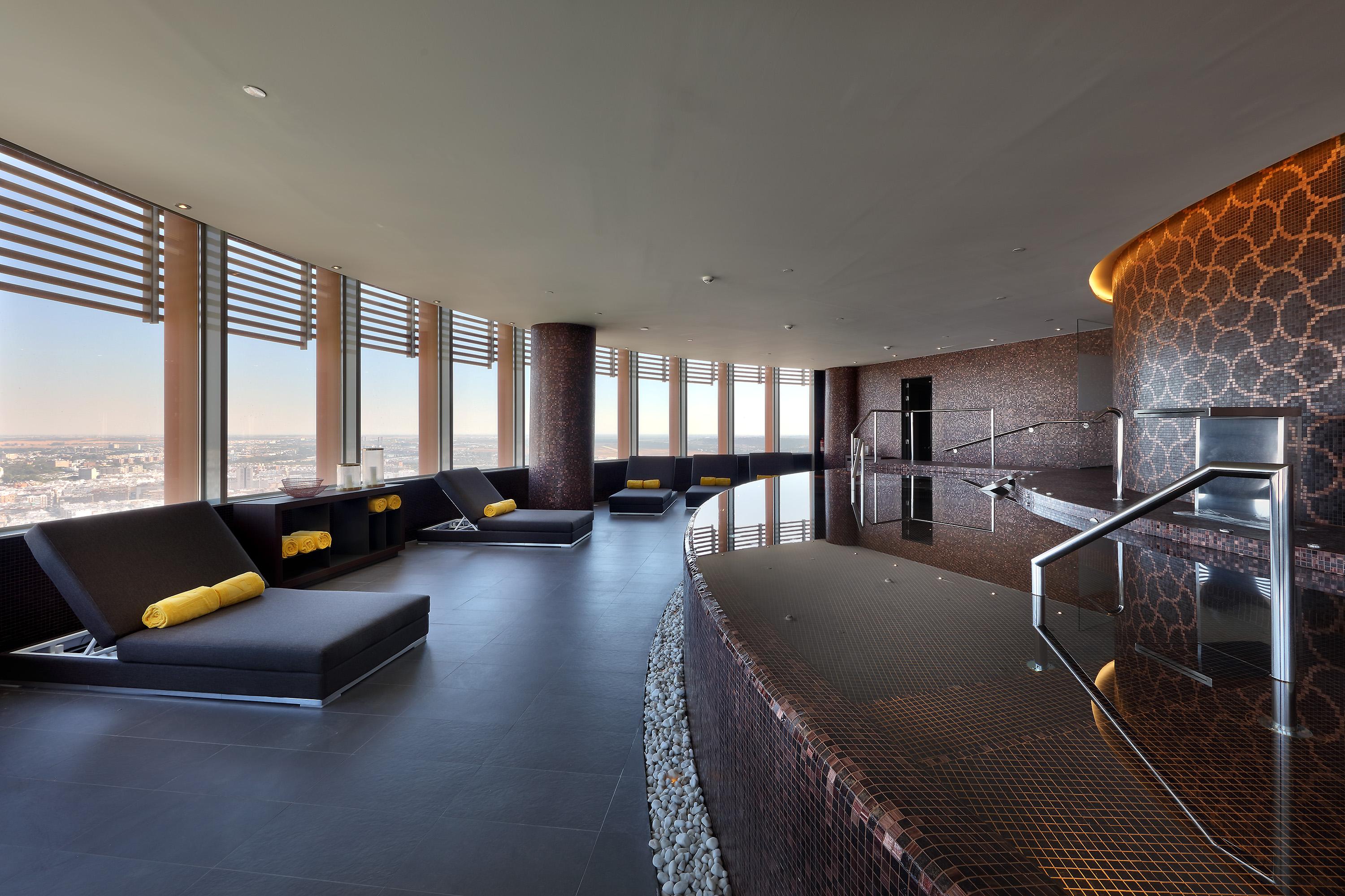 Eurostars Torre Sevilla Exterior foto The photo depicts a modern and elegant spa interior. It features a curved layout with large windows providing a panoramic view. The space includes several lounge chairs with yellow cushions, arranged in a relaxed manner. A sleek, dark-finished counte