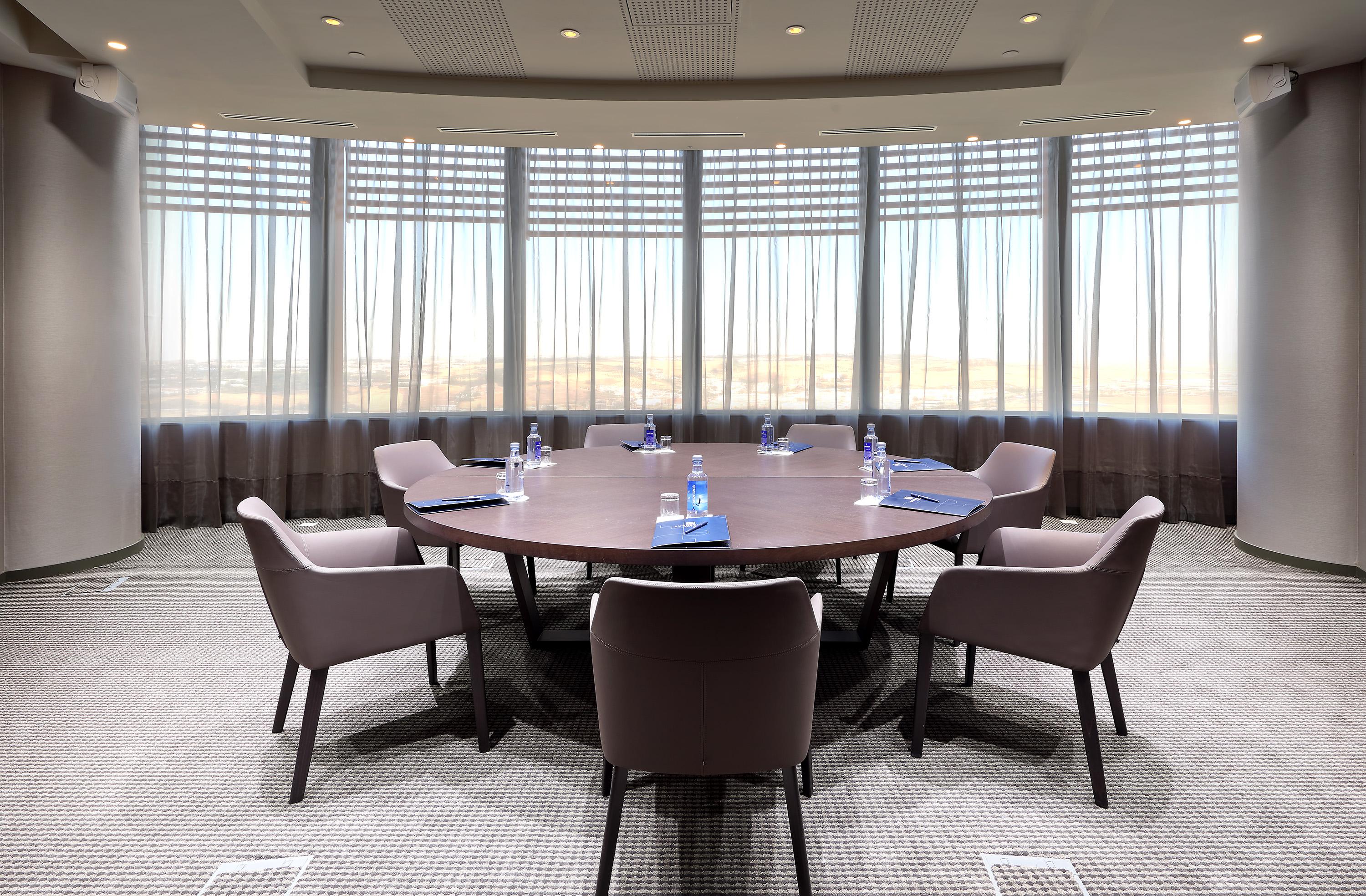 Eurostars Torre Sevilla Exterior foto The photo shows a conference room with a large round table at its center. The table is surrounded by several modern chairs. There are water bottles placed at each seat, and blue notepads or documents are also visible on the table. The room has large 