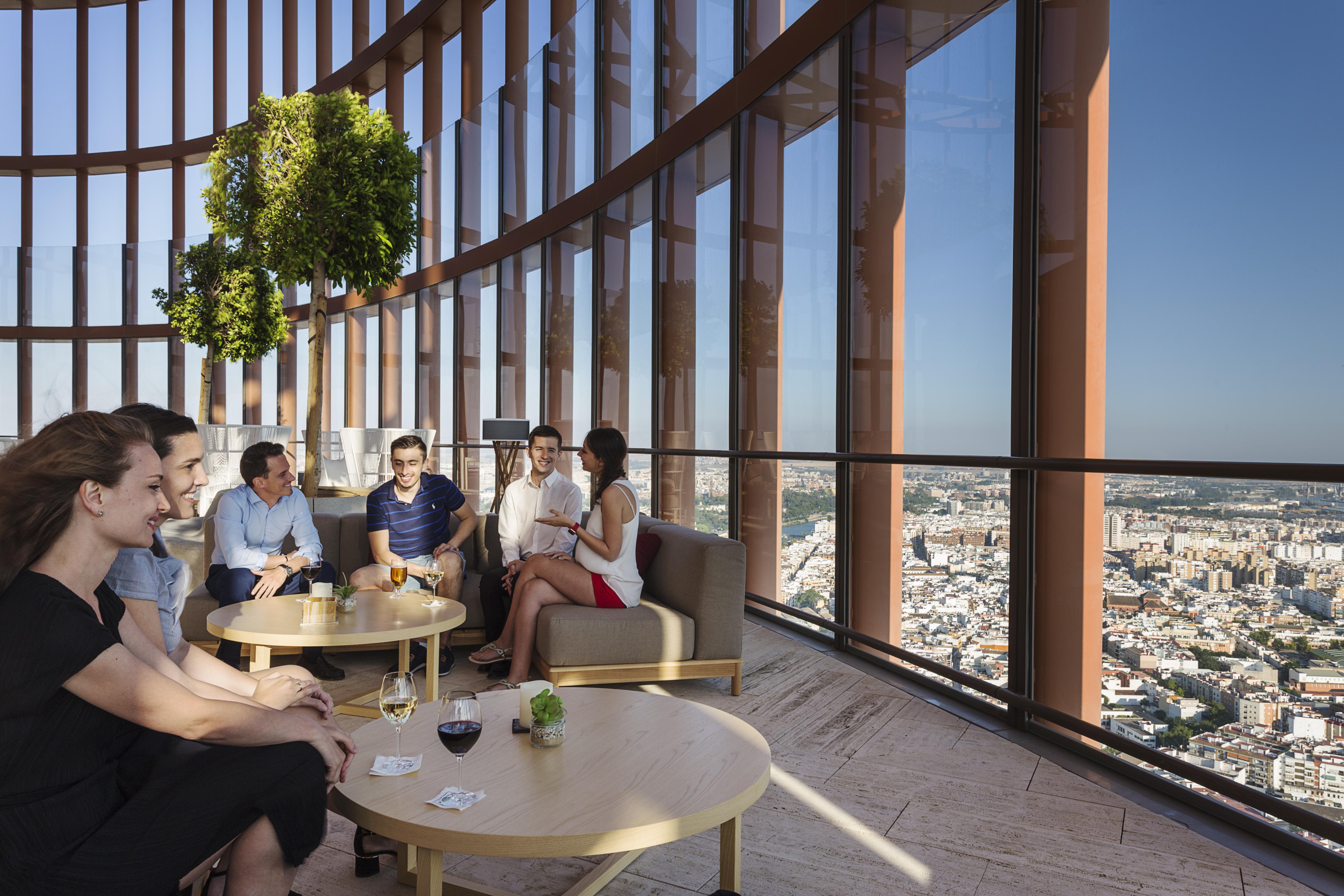 Eurostars Torre Sevilla Exterior foto The photo shows a group of people sitting in a modern, stylish lounge area with large windows that provide a panoramic view of a cityscape below. The setting appears to be high up in a building, possibly a skyscraper. There are several tables with dr