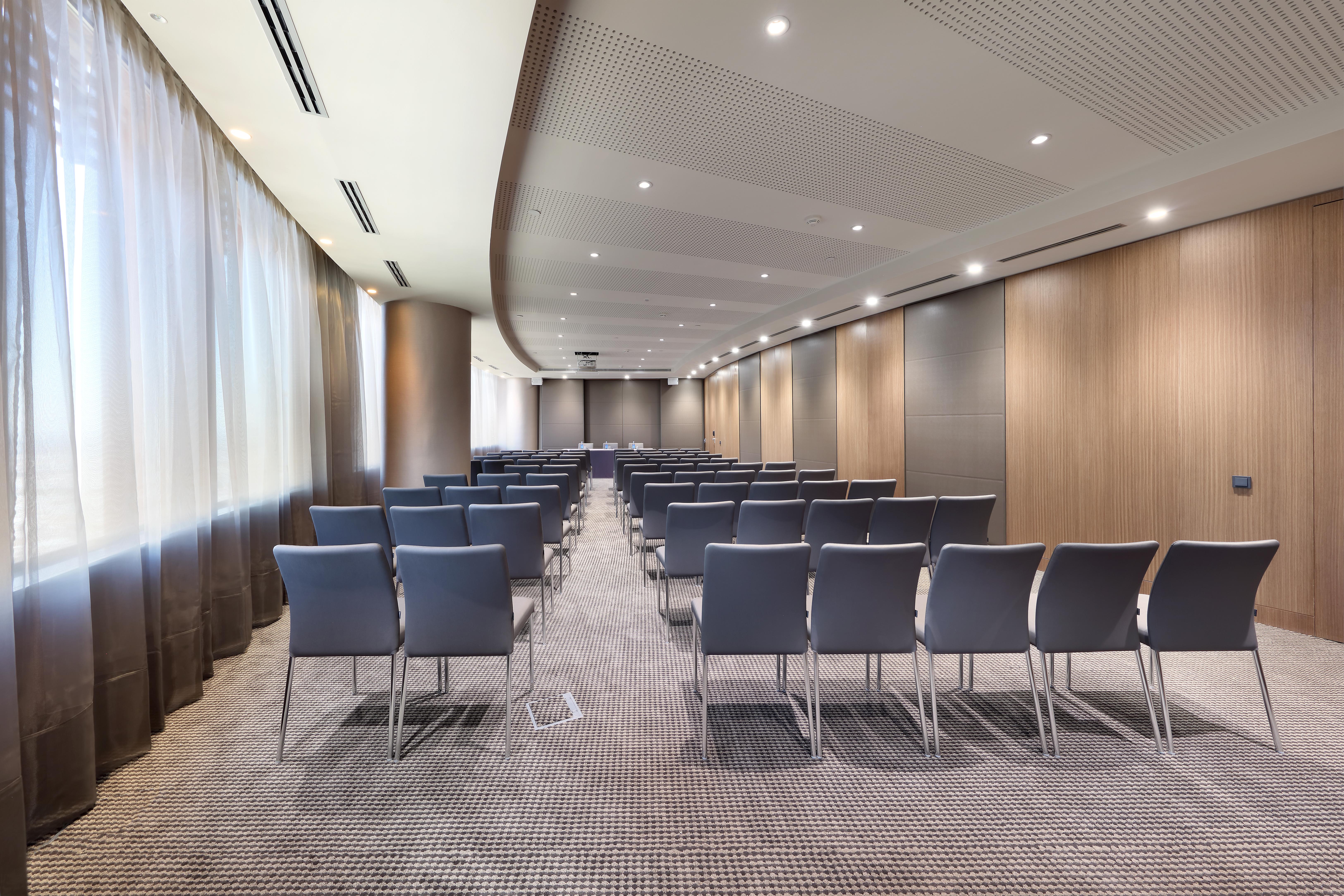 Eurostars Torre Sevilla Exterior foto The photo shows a modern conference room with an elongated layout. There are rows of grey chairs arranged neatly facing a focal point at the front of the room, likely for a presentation or meeting. The walls are a combination of wooden paneling and n
