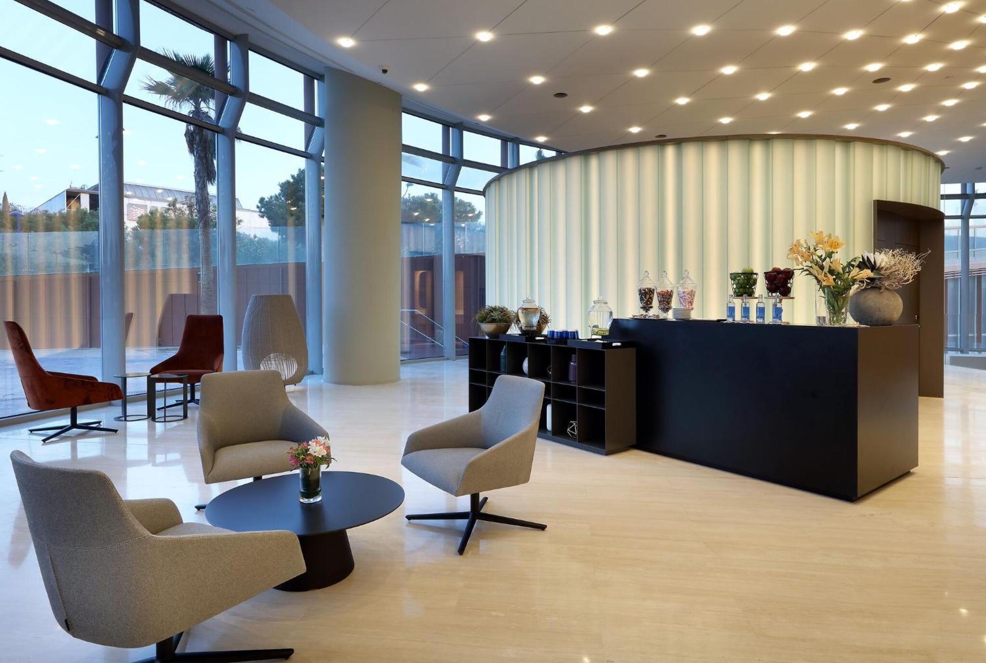 Eurostars Torre Sevilla Exterior foto The photo depicts a modern reception area. There is a sleek, rounded reception desk in the center against a light-colored wall with vertical stripes. Behind the desk, various decorative items and flowers are displayed. In front of the desk, there are