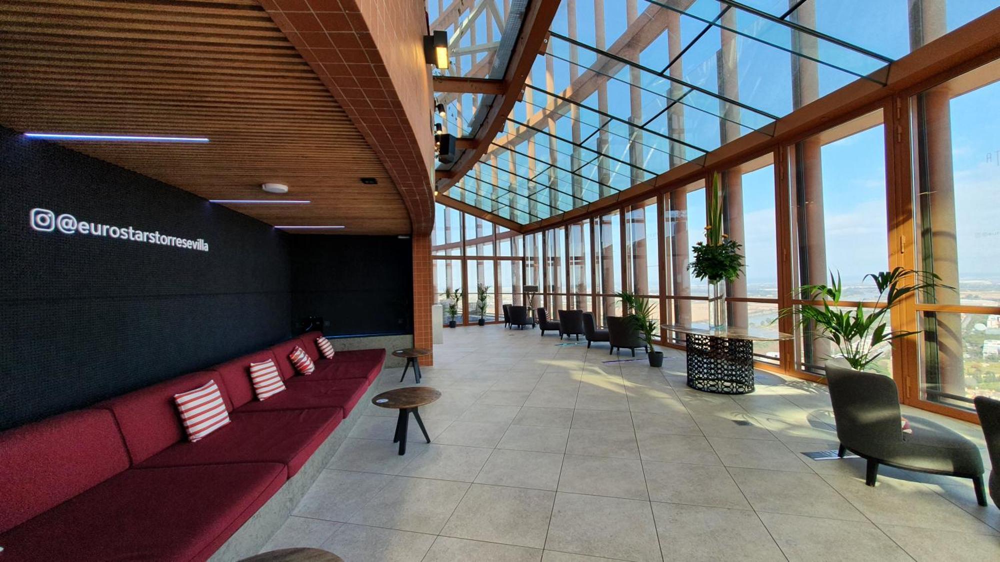 Eurostars Torre Sevilla Exterior foto The photo shows a spacious, modern interior with a large glass wall and ceiling that allows for ample natural light. The area features a long red couch with several cushions, paired with small tables. There are potted plants throughout the space, add