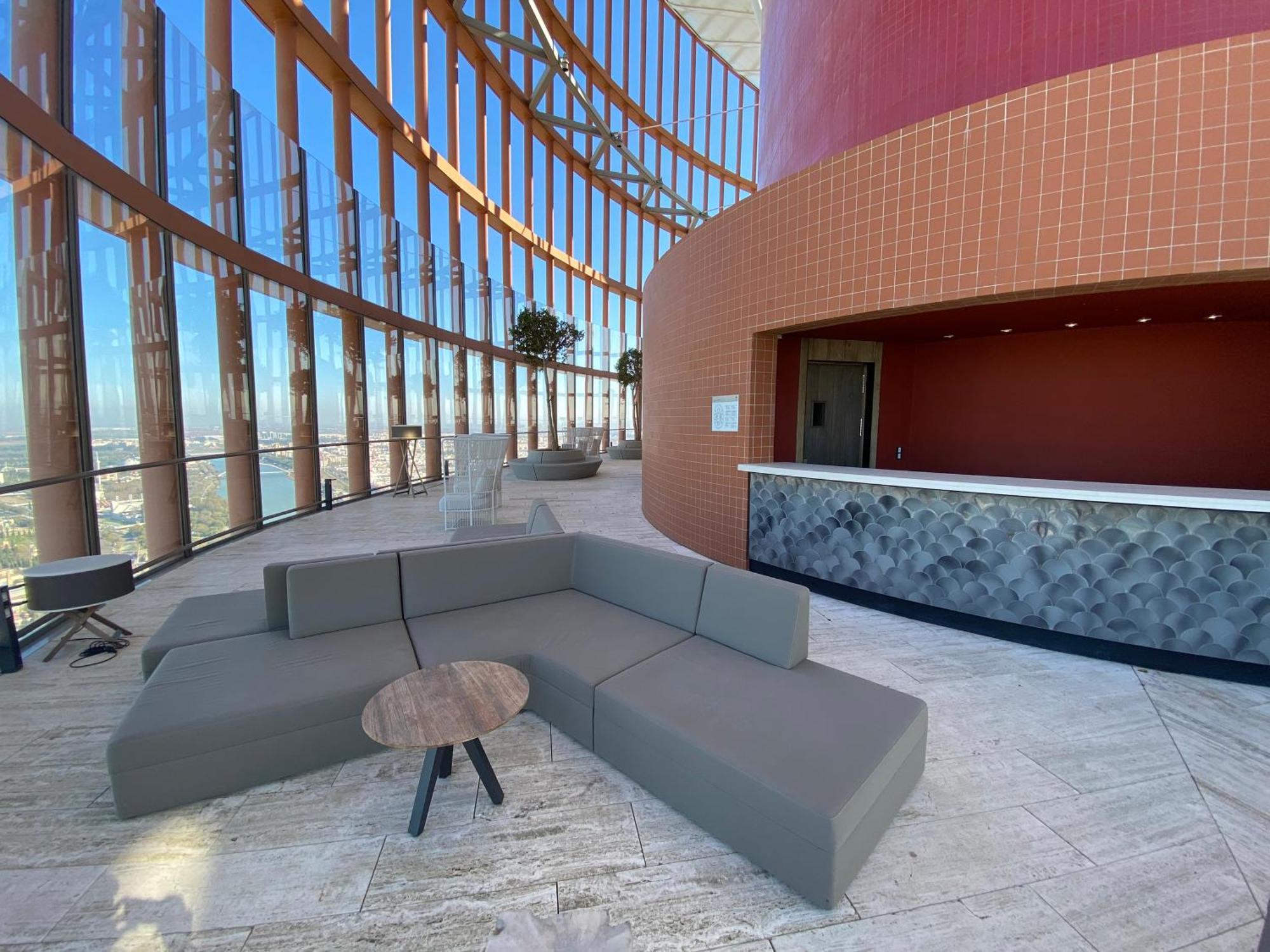 Eurostars Torre Sevilla Exterior foto The photo shows a modern interior space with large windows that provide a panoramic view. There is a comfortable, modular gray seating arrangement shaped like a curved couch, paired with a small, round table. In the background, there's a reception or