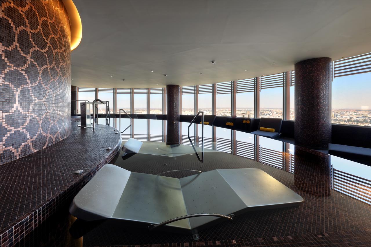 Eurostars Torre Sevilla Exterior foto The photo depicts a modern spa or wellness area with large windows offering a panoramic view of the surroundings. In the foreground, there are two sculptural lounge areas that appear to be partially submerged in water. The interior features elegant t