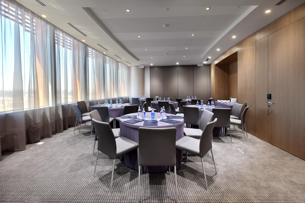Eurostars Torre Sevilla Exterior foto The photo shows a modern conference room featuring several round tables arranged in the center. Each table is covered with a purple tablecloth and has bottles of water set on top. There are gray chairs positioned around the tables. The room has large