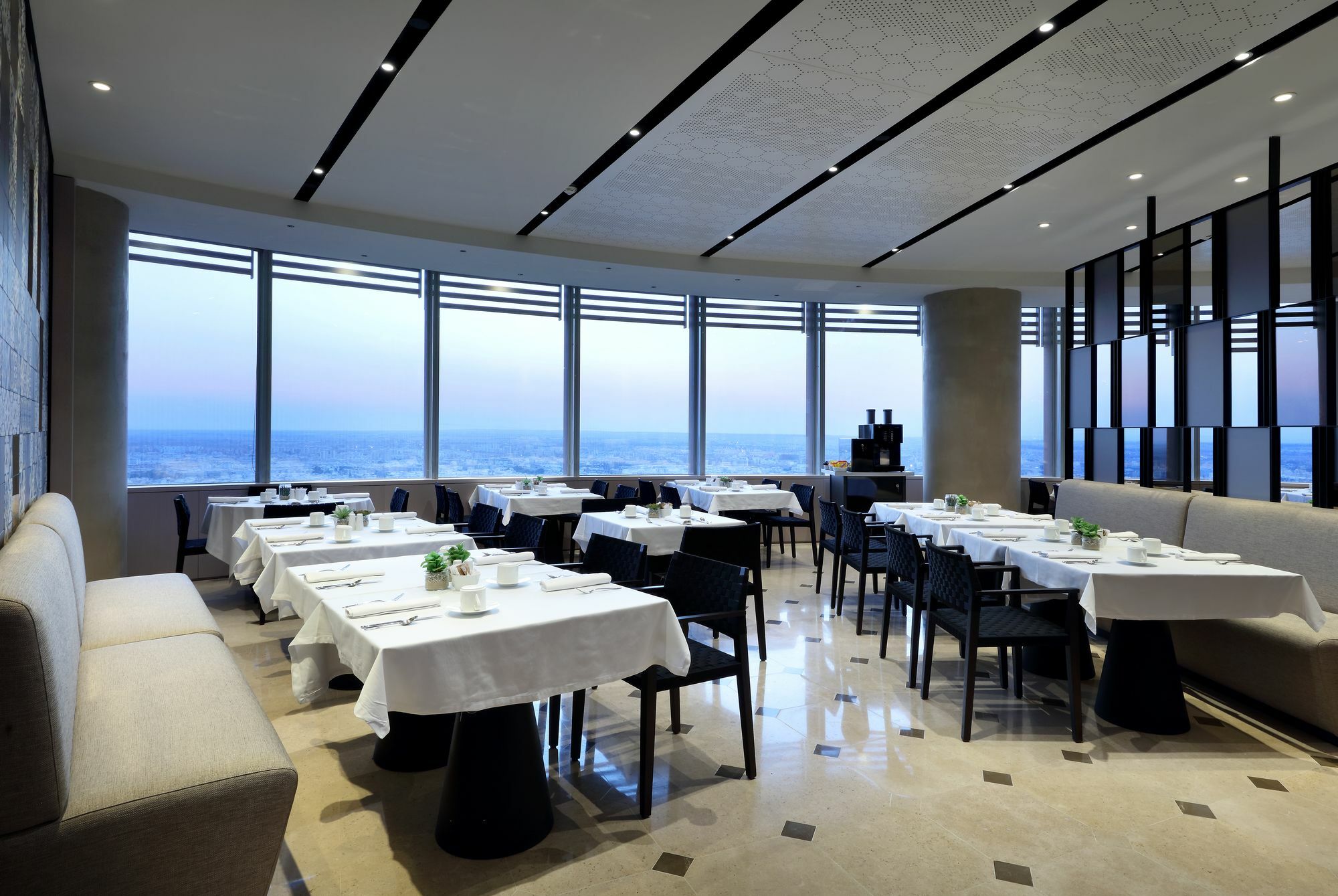 Eurostars Torre Sevilla Exterior foto The photo shows the interior of a modern restaurant. The space features multiple dining tables arranged in an elegant layout, each covered with white tablecloths. The chairs are sleek and dark, complementing the contemporary design. Large windows lin