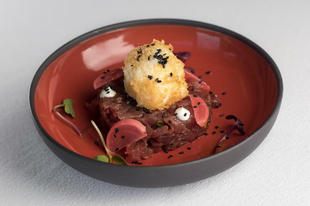 Eurostars Torre Sevilla Exterior foto The photo shows a dish presented in a round, orange bowl. The main component appears to be a tartare, possibly made of raw meat, finely chopped and garnished with small pieces of what looks like radish. Atop the tartare, there's a round, golden-brown
